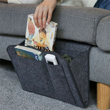 Ein graues BedCaddy mit zwei Büchern und einer Fernbedienung hängt an der Seite einer grauen Couch.