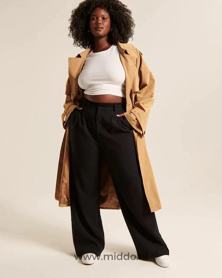 Eine dunkelhäutige Dame mit einem langen beigefarbenen Mantel präsentiert die schwarzeweite Hose mit weißen Sportschuhen darunter.